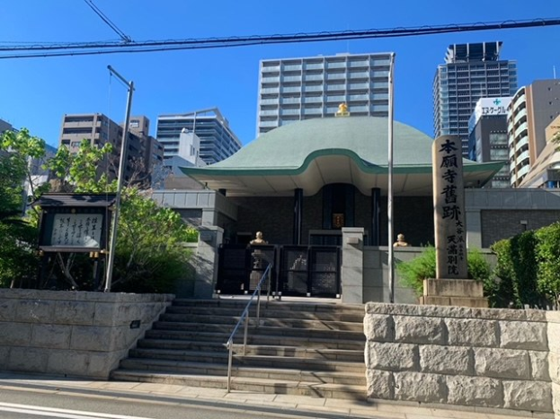 Großes grünoffenes Gebäude in städtischer Umgebung mit umliegenden Treppen, Geländern, Beschilderung, Grünflächen, einer Straße unten und Wolkenkratzern im Hintergrund unter einem klaren blauen Himmel.
